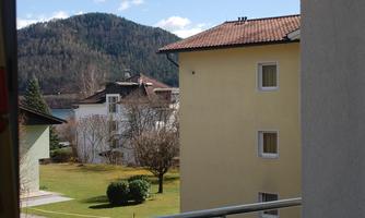 A balcony view of a green meadow and surrounding houses. In the background, gentle hills and a blue sky with white clouds can be seen.