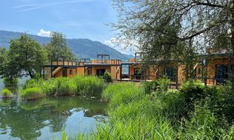 A modern holiday home by the shore of a pond, surrounded by lush greenery. In the background, mountains and a blue sky can be seen.