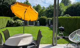 A cozy garden with a large, round table and a yellow umbrella. In the background, beautiful hedges and a grill can be seen.