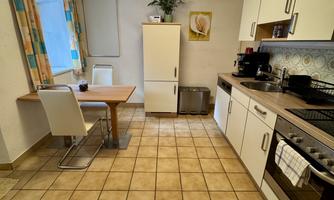 A bright kitchen with modern furniture and a table. Large windows let in plenty of light, and the tiles are laid out in a practical pattern.