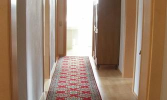 A bright hallway with a colorful carpet and wooden frames on the doors. At the end of the hallway, there is daylight and a wardrobe.