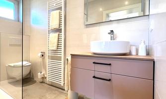 A modern bathroom with a glass shower cabin and light tiles. The sink area features a stylish wooden dresser and a large mirror.