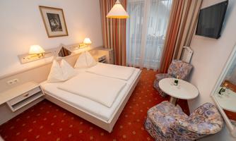 A simple room with a double bed, two chairs, and a small table. The room has light walls and a television on the wall.