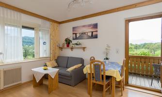 A bright living room with a cozy sofa and a dining area. Large windows offer a view of the green landscape outside.
