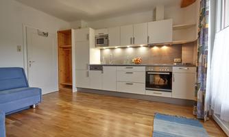 A modern kitchen with bright furniture and a cozy living room. The floor is made of wood and there is plenty of light from the windows.