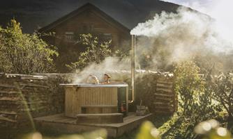 A wooden hot tub in a picturesque garden landscape. Steam is rising, and a wooden house can be seen in the background.