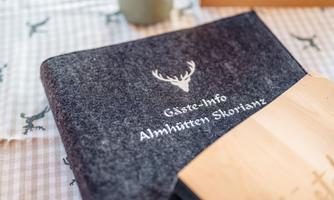 A dark gray guestbook with the imprint "Guest Info Almhütten Skorianz". In the background, there is a table with a checked pattern.