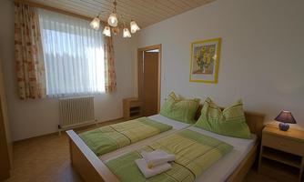 A cozy bedroom with two beds and colorful cushions. The room is bright and inviting, with a window and a decorative lamp.