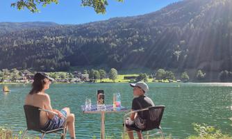 Two people are sitting at a table on the shore of a clear lake. In the background, green hills and mountains stretch out.