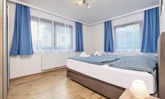 A modern bedroom with a large bed, blue curtains, and light wood flooring. There is a relaxed atmosphere with natural light.