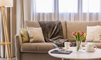A cozy living room with a bright sofa, decorative cushions, and a warm blanket. On the table, there are flowers, a cup, and papers.
