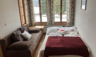 A cozy room with a bed and a couch. Large windows allow plenty of daylight in and provide a view of the surroundings.