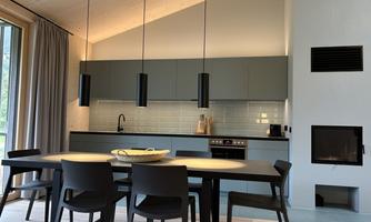 A modern kitchen with a large dining table and black chairs. Ceiling lights hang above the table and there is a fireplace in the corner.