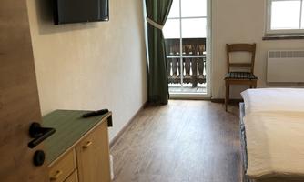 A simple bedroom with wooden flooring and a large window. A television and a small desk are also available.
