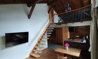 A cozy living space with a wooden beam ceiling and a staircase. A dining table with flowers and a television on the wall are also visible.