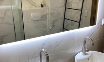 A modern bathroom with two sinks and extensive mirror lighting. In the background, a shower with a glass wall is visible.