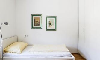 A simple bedroom with a single bed and a reading lamp. Two framed pictures hang on the wall.
