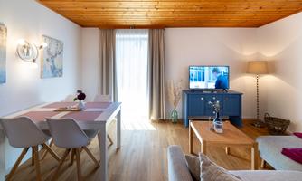 A modern living room with a wooden ceiling and cozy furnishings. There is a table with chairs, a sofa, and a television in front of a window.