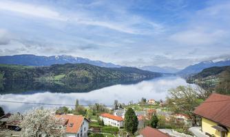 A picturesque view of a lake, surrounded by mountains and wooded slopes. The clouds gently reflect in the water.