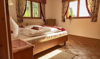 A bright bedroom with a double bed and a wooden floor. The windows let in plenty of daylight, and the room is cozily decorated.