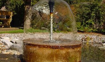 A water fountain bubbles from a round stone basin. The surroundings are green and natural, with trees in the background.