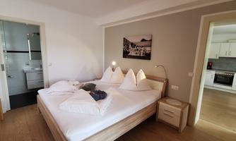 A cozy bedroom with a large bed and white bedding. In the background, a bathroom and a kitchen are visible.