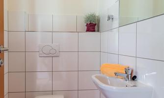 A modern bathroom with white tiles and a small window. There is a sink, a toilet, and a plant on the windowsill.