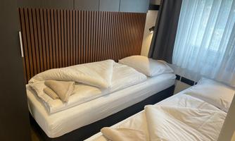 A modern bedroom with two single beds and wood paneling. The windows are decorated with light curtains, allowing plenty of daylight in.