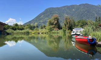 A calm lake surrounded by green trees and mountains. A red boat floats at the shore.
