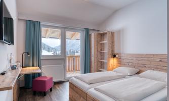 A modern bedroom with two single beds and a large window. The wooden furniture and the window front provide a beautiful view of the mountains.