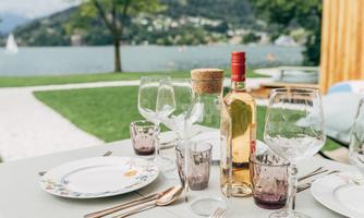 A beautifully set table with glasses, cutlery, and a bottle of wine is located by the shore of a lake. In the background, green hills and a clear sky can be seen.