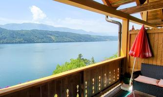 A beautiful view of a calm lake from a wooden terrace. In the background, mountains and a clear sky are visible.