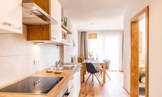 A modern kitchen with wooden shelves and a clear workspace. The adjoining dining area is bright and inviting, with a table and chairs.
