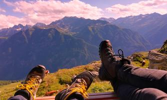 Two legs in hiking boots enjoy the view of the mountains. In the background, a beautiful mountain landscape with green grass and clouds in the sky can be seen.