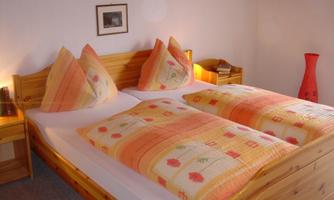 A cozy bedroom with two beds and colorful cushions. The walls are light and a red decorative pot stands in the corner.