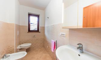 A modern bathroom with bright tiles and a large window. There is a sink, a toilet, and a bidet, as well as a wooden cabinet.
