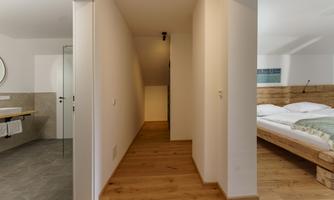 A bright hallway with wooden flooring and white walls. On the left side is a bathroom and on the right is a cozy bedroom with a wooden bed.