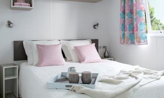 A bright bedroom with a large bed and pink pillows. On the bed, there is a tray with two cups and a blanket.