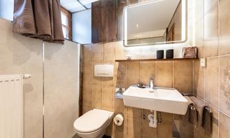 A modern bathroom with bright wall mirror lighting. The tiles are in warm earth tones, and there is a shower as well as basic sanitary facilities.