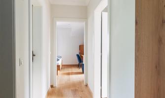 A bright hallway with wooden flooring and several doors. At the end of the hallway, a room with furniture can be seen.