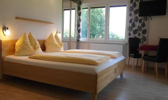 A cozy bedroom with a large bed and yellow pillows. There are large windows with a view of greenery and a table with chairs.