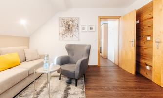 A modern living room with a gray armchair and a beige couch. The floor is made of wood, and there is a table with water and decorative wall art.