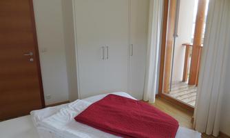 A bright bedroom with a white bed and a red blanket. Behind the bed are large windows and a door leading to a balcony.