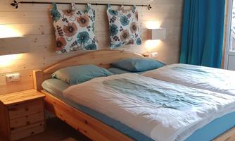 A cozy bedroom with a large double bed and bright, colorful pillows. The walls are made of wood, and there is a beautiful lamp as well as blue curtains.
