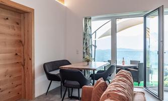 A modern living room with a dining table and black chairs. Large windows provide a view of the landscape and plenty of daylight.