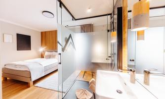 A modern hotel room with a large bed and wooden details. The bathroom is open-plan and features an elegant sink area.