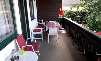 A cozy balcony with white furniture and a red table. In the background, green plants and a sunshade can be seen.