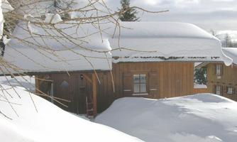 A wooden house is surrounded by a thick blanket of snow. The sky is cloudy and the landscape is wintery and quiet.