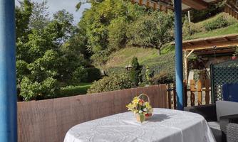 A table with a tablecloth is on a terrace, surrounded by green garden. In the background, there are trees and gentle hills.