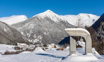 A wintry landscape with snow-covered mountains and a small village in the background. In the foreground, there is a modern statue in a snowy field.